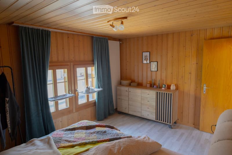 Cozy wooden bedroom with a bed, a cat hammock, a wooden cabinet, and a radiator. The room features wooden walls and ceiling, large windows with dark curtains, and a small cat sitting on the hammock.
