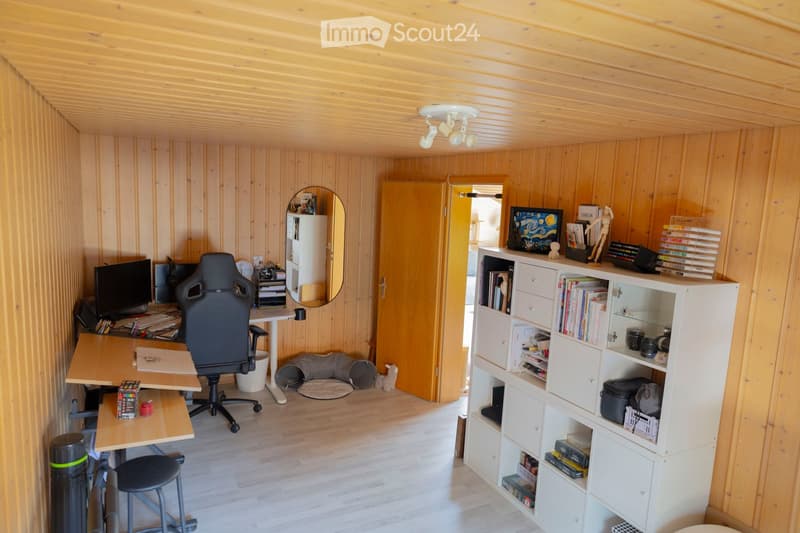 A cozy, wooden-paneled room with a desk setup, a bookshelf, and a small seating area. The room features a wooden ceiling and walls, a large mirror, and a small dog bed in the corner.