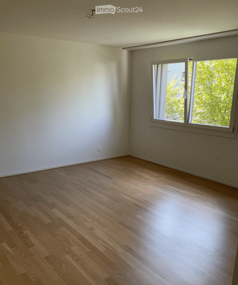 Empty room with wooden flooring and large windows allowing natural light. Walls are painted white, and there are no furnishings or decorations present.
