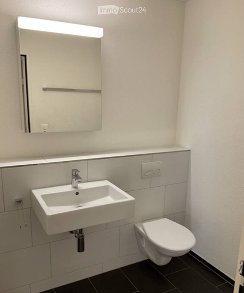 Modern bathroom with a white sink, toilet, and a large mirror with built-in lighting above it. The walls are tiled in white and grey, and the floor is black.