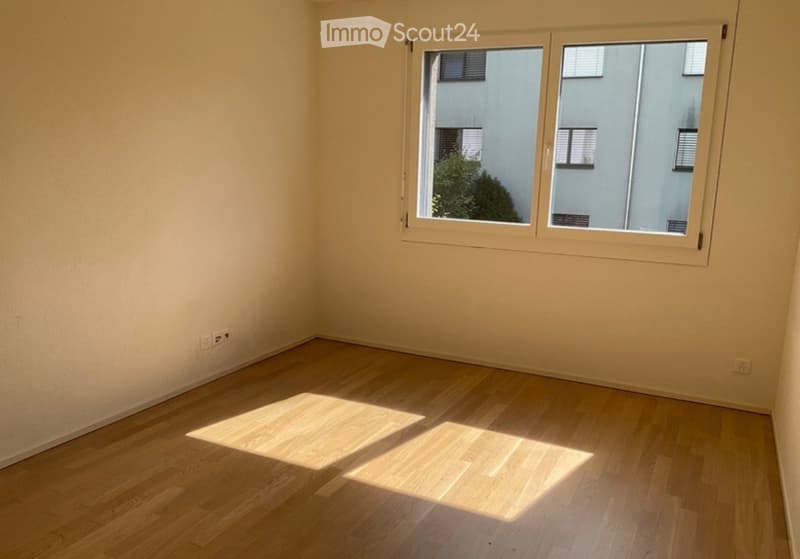 Empty room with light wooden flooring and large windows allowing natural light to enter.