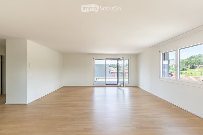 Empty room with light wooden flooring, white walls, large windows, and a set of sliding glass doors leading to a balcony.