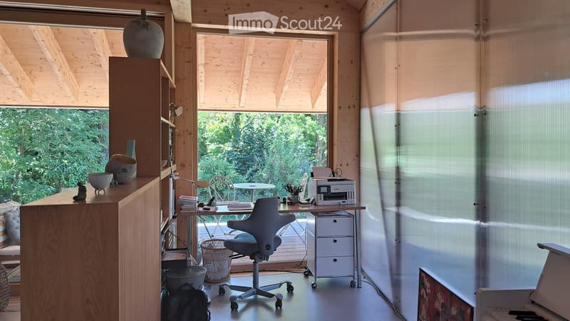Bright, modern workspace with wooden walls and ceiling, large windows, and a view of greenery. Features a desk with a chair, printer, and various office supplies, along with a side table and decorative items.