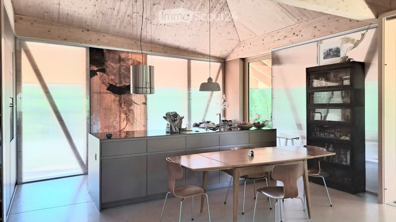 Modern kitchen with grey cabinets, a central island, a wooden dining table with four chairs, pendant lights, and a large window offering a view of the garden.