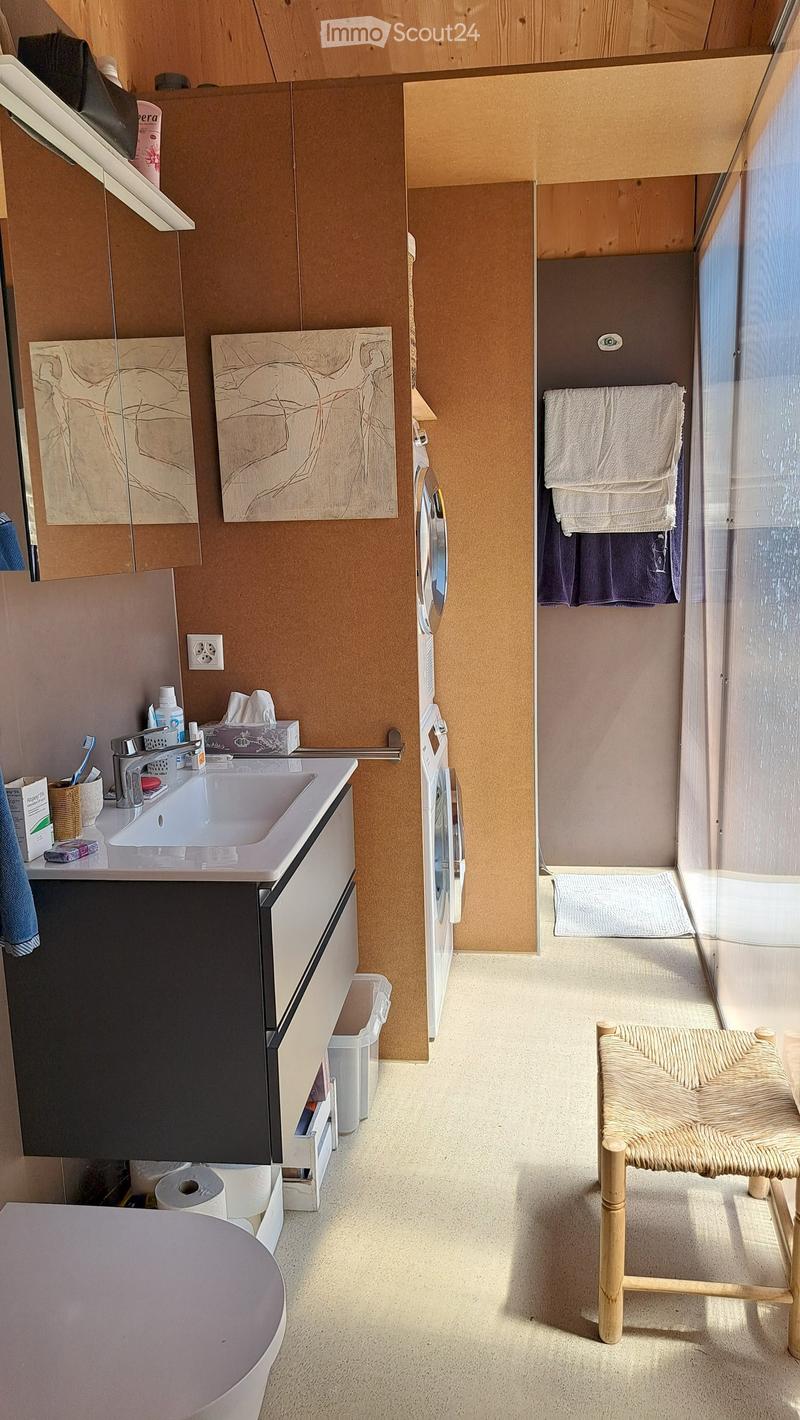 A small, modern bathroom with a sink, mirror, and a washing machine. A wicker chair is placed near the shower area, and towels are neatly arranged on a rack.