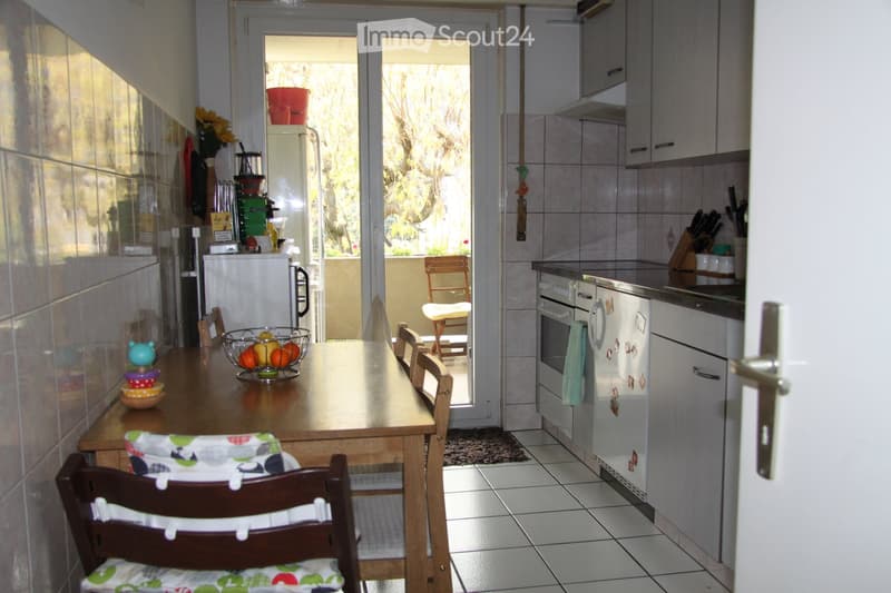 A modern kitchen with white cabinets, a wooden dining table, and various appliances including a refrigerator, oven, and dishwasher. The room features tiled floors and walls, with a view to a balcony through large glass doors.