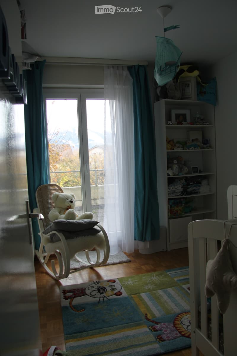Brightly lit children's bedroom with wooden flooring, a rocking chair with a teddy bear, a colorful rug with animal prints, and a shelf filled with toys and books.
