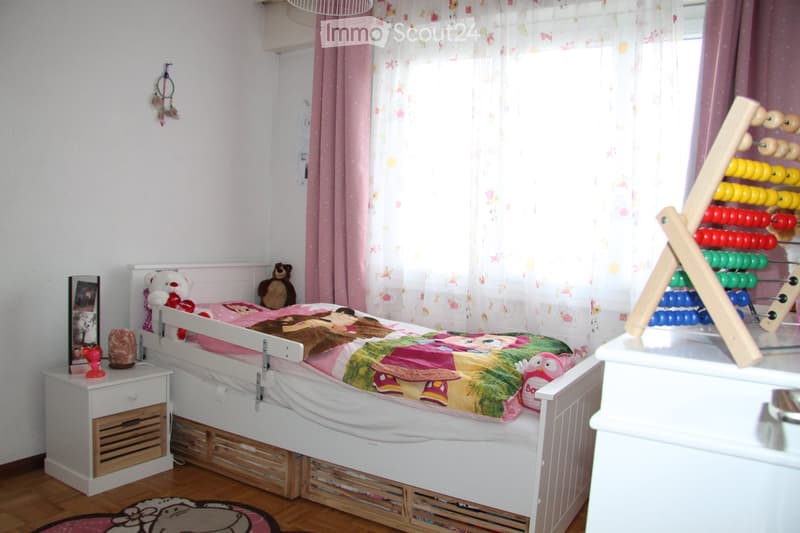A child’s bedroom with a white bed, colorful quilt, nightstand, and various toys including an abacus and stuffed animals.