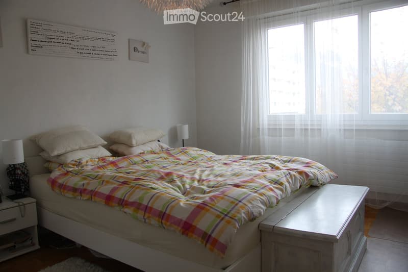 A cozy bedroom with a neatly made bed, colorful plaid duvet, nightstands with lamps, and sheer curtains on the windows.
