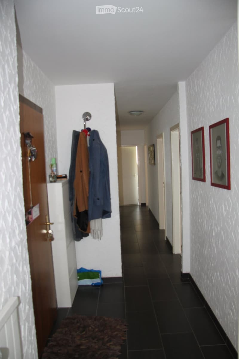 A narrow hallway with white walls, dark tiled flooring, and a coat rack holding jackets. There are framed pictures on the right wall and a small rug on the floor.