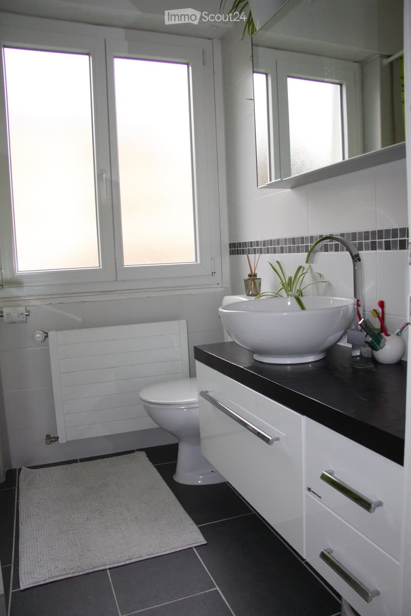 Modern bathroom with white tiles, black countertop, white sink, chrome faucet, toilet, and a small plant on the counter.
