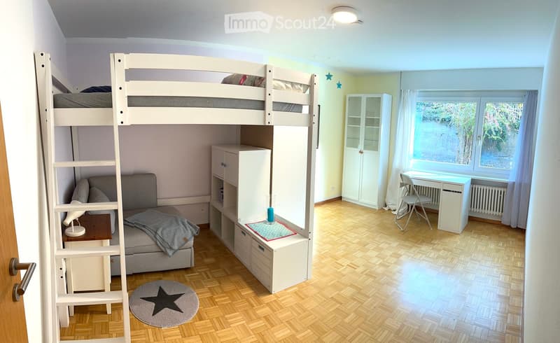 A well-lit bedroom with a bunk bed, a small sofa, a desk, and a chair. The room features wooden flooring, white walls, and large windows with curtains.