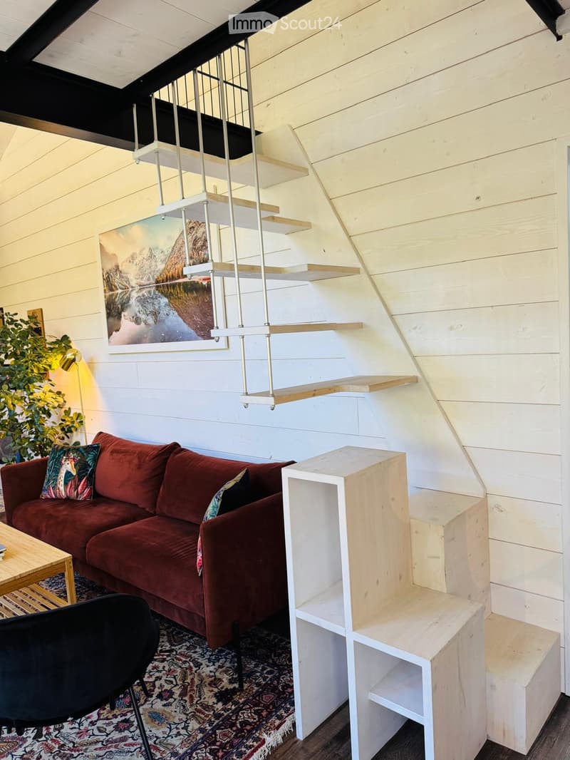 A cozy living room with a red sofa, wooden coffee table, black chair, and a staircase leading to a loft area. The room features wooden walls, a patterned rug, and a painting on the wall.