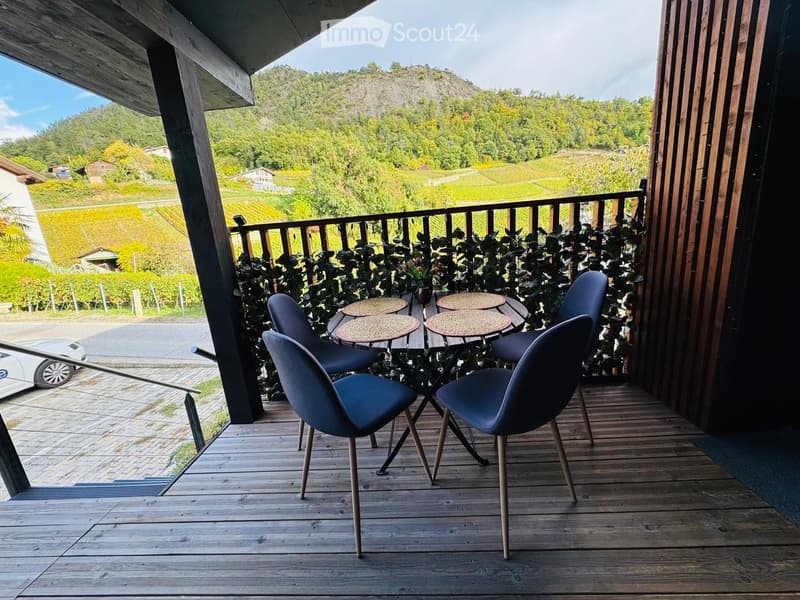 Balcony with wooden decking, small round table, four blue chairs, and a scenic view of vineyards and hills.