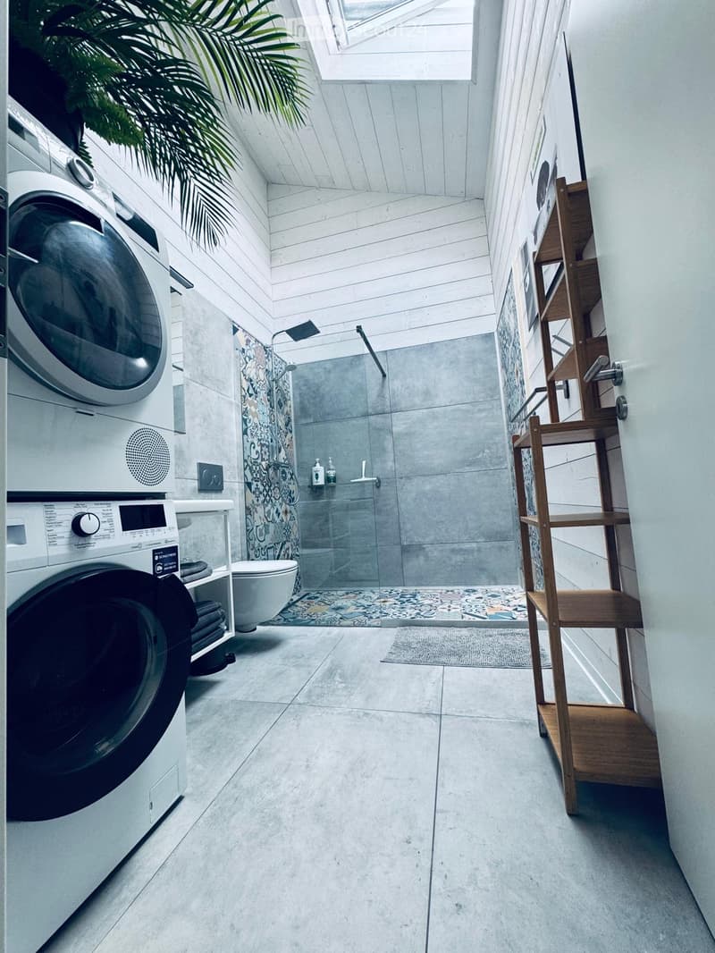 Modern bathroom with a washing machine, toilet, wooden shelf, and a glass-enclosed shower area with a decorative shower curtain.