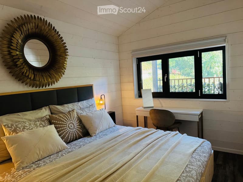 Cozy bedroom with a wooden bed, patterned pillows, a large mirror with a sun-shaped frame, a small desk with a chair, and a window with a view of greenery.