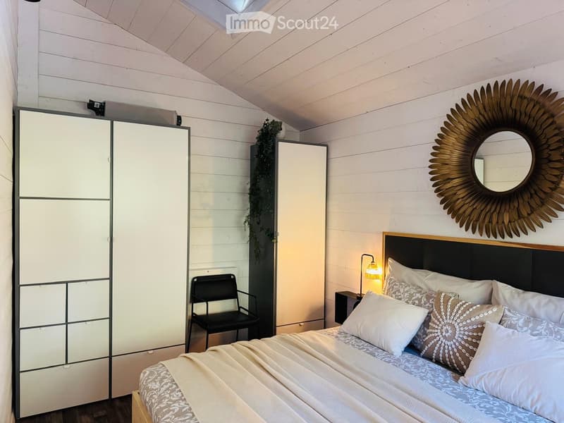 Cozy bedroom with a large bed, patterned bedding, a modern wardrobe, a black chair, and a decorative mirror on the wall.