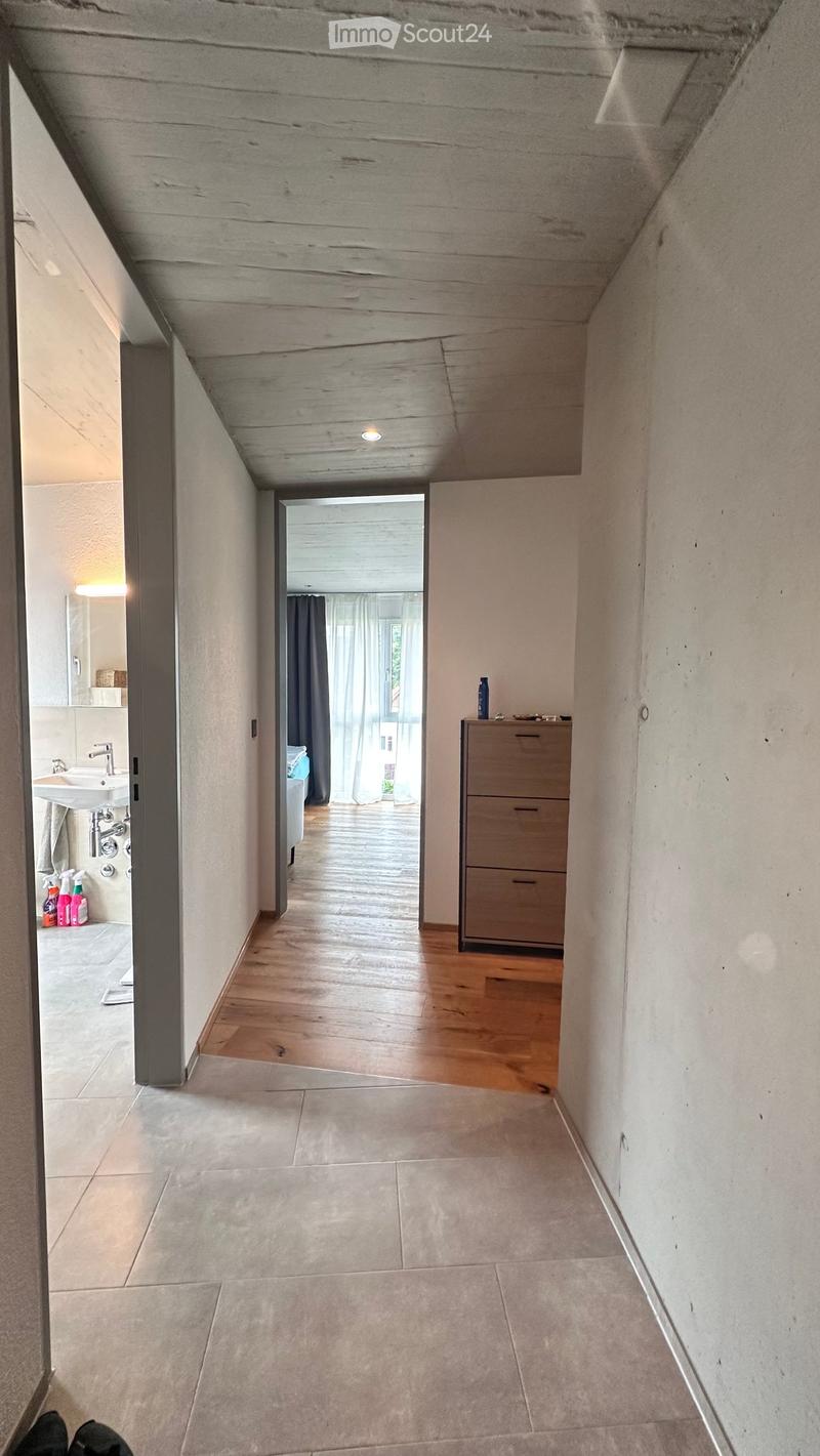 Modern hallway with exposed concrete ceiling, wooden flooring, and a small cabinet with a bottle on top. A bathroom is visible to the left, and a room with a window and curtains is visible ahead.