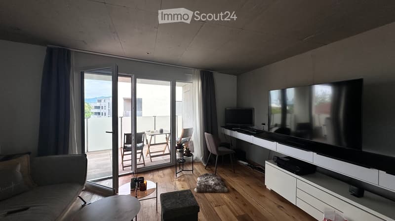 Modern living room with a grey sofa, coffee table, round side table, and a large TV screen on a white cabinet. There is a workspace with a laptop and chair, and a small table and chairs on a balcony accessible through large glass doors.