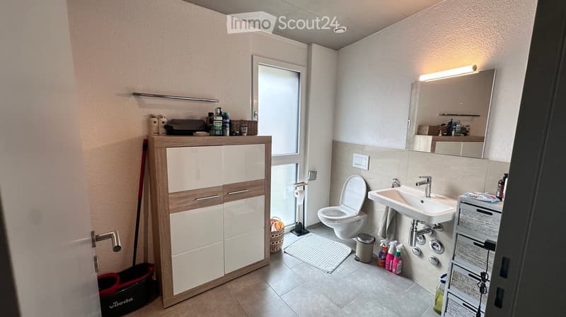 Modern bathroom with white cabinetry, a sink, toilet, large mirror, and cleaning supplies neatly arranged. Features a window for natural light and a small basket on the floor.
