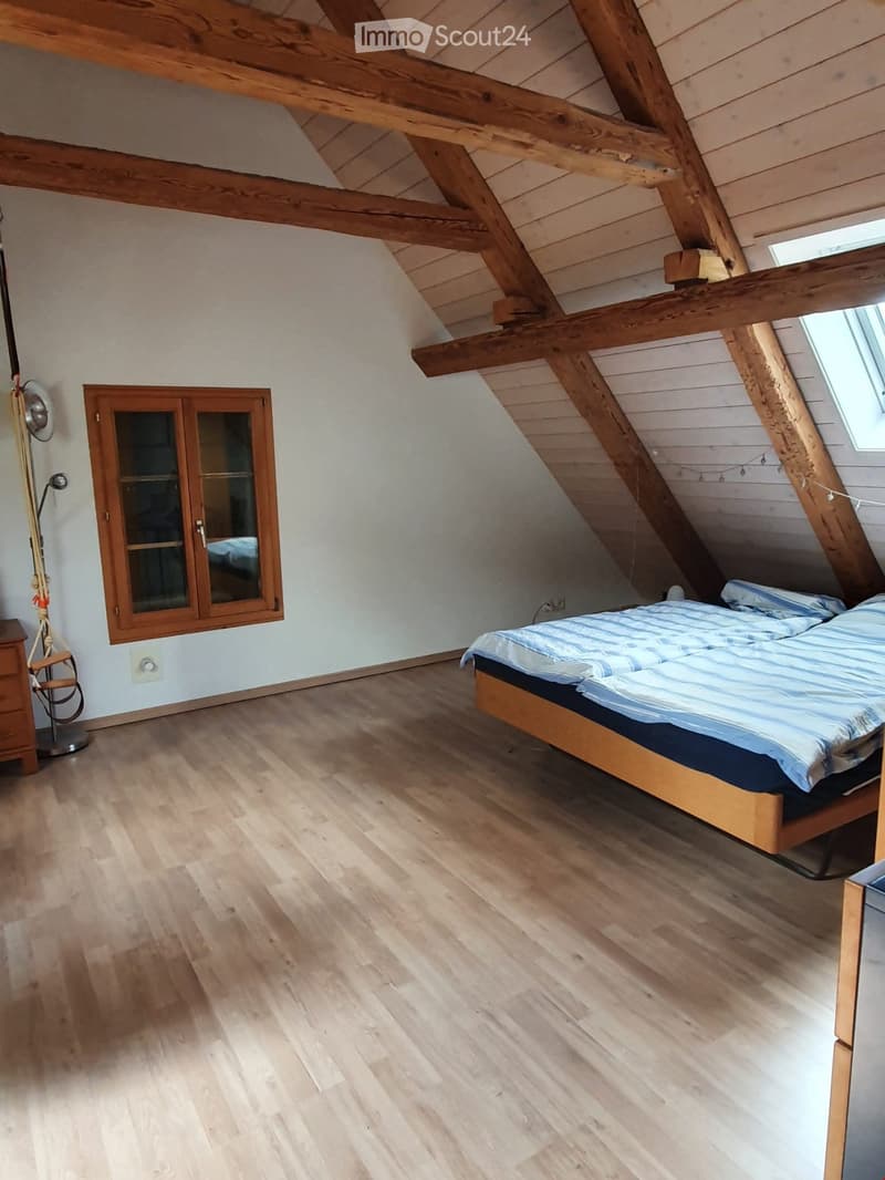 Bright attic bedroom with wooden beams, light wooden flooring, a double bed with blue and white bedding, a small wardrobe with glass doors, and a standing fan near the wall.