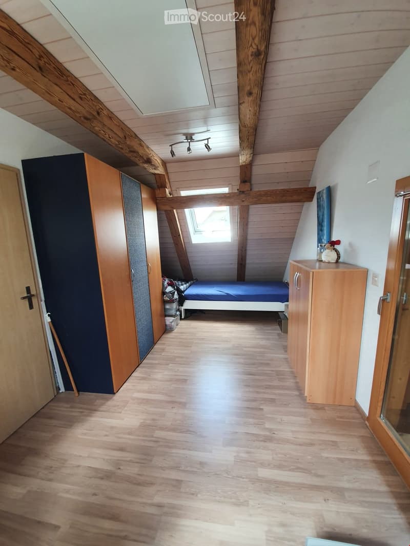 Cozy attic bedroom with wooden floors, slanted wooden ceiling beams, and built-in storage. Features a single bed, wardrobe, and small cabinet with decorative items.