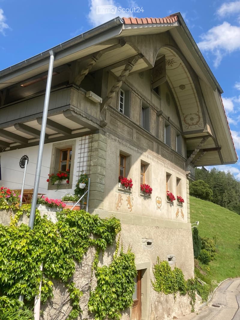 A multi-story stone house with decorative carvings, balconies, and flower boxes filled with red and pink flowers. The building features a tiled roof and is surrounded by greenery and a grassy hillside.