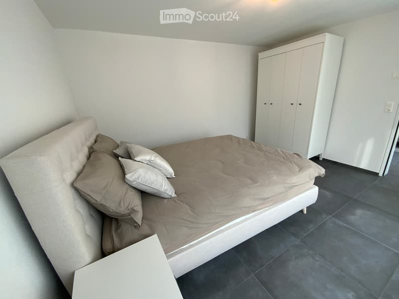 Modern bedroom with a white bed, beige bedding, and a white wardrobe. The room features dark grey tiled flooring and white walls.