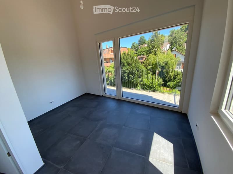 Empty room with dark grey tiled floor, white walls, large sliding glass door leading to a balcony with a view of greenery and neighboring houses.