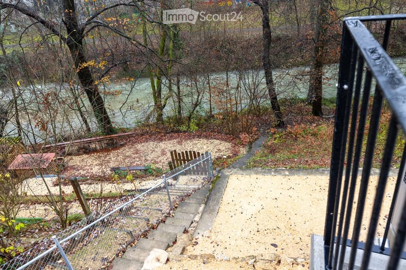 Outdoor view from a balcony showing a staircase leading down to a garden area with trees, a river in the background, and a small wooden structure.