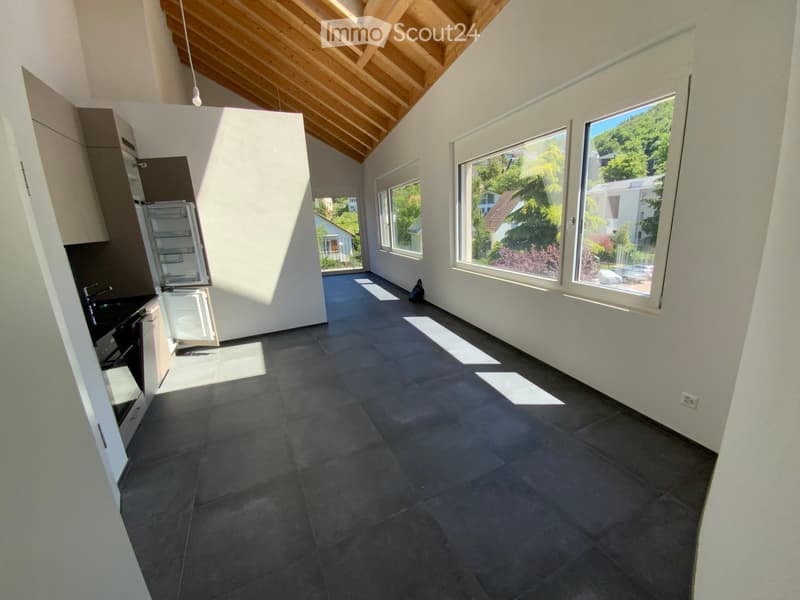 Open-plan space with a modern kitchen area on the left and large windows on the right, featuring a dark tiled floor and white walls.