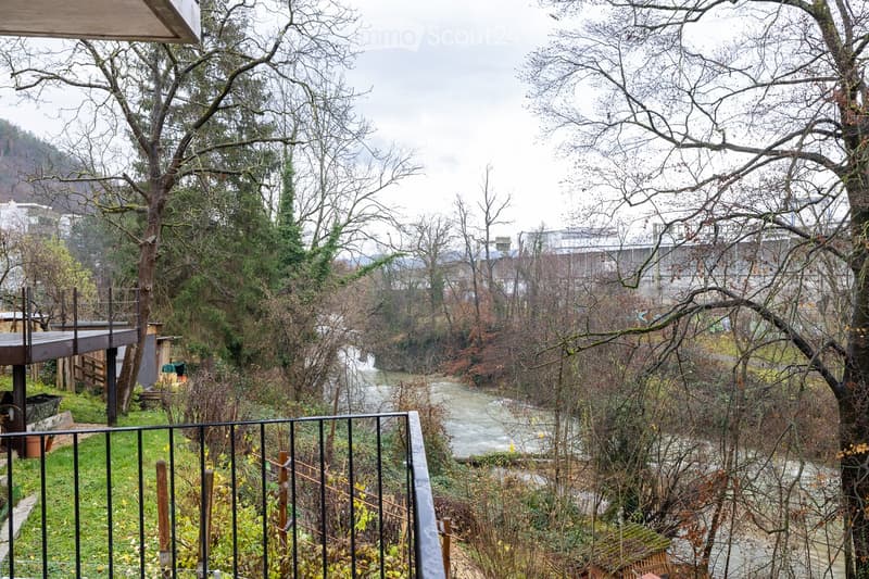 Balcony overlooking a river with trees and a wooden deck visible in the background.