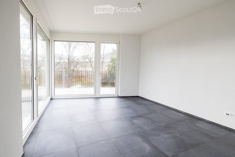 Empty room with white walls, grey tiled floor, large sliding glass doors leading to an outdoor area, and a metal fence visible outside.