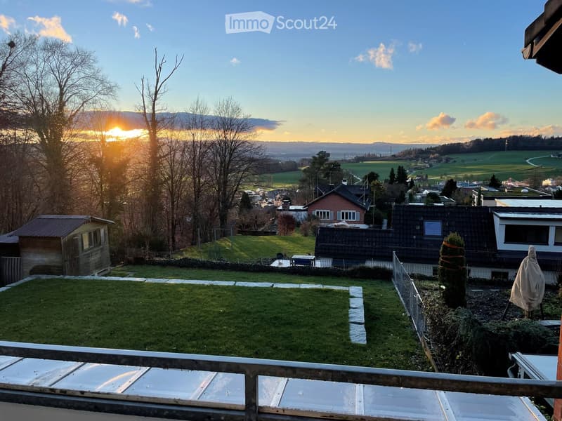 View from a balcony or terrace showing a grassy yard, surrounding houses, trees, and a scenic sunset view of hills and fields.