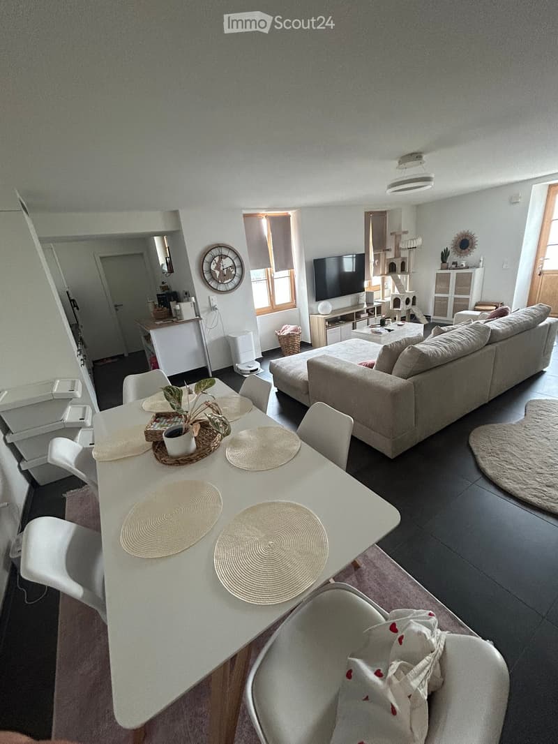 Modern living room with a grey sofa, coffee table, cat tree, dining table with chairs, and a television mounted on the wall. The room features a large window with blinds and a small kitchen area visible in the background.