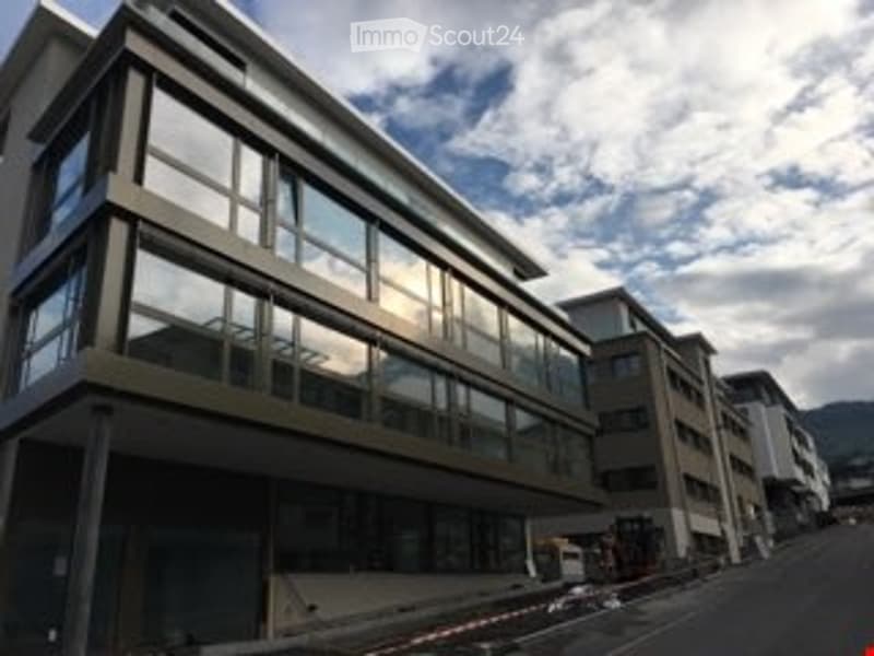 Modern multi-story building with large glass windows and a sleek design, situated in an urban area with a clear sky and distant mountains.
