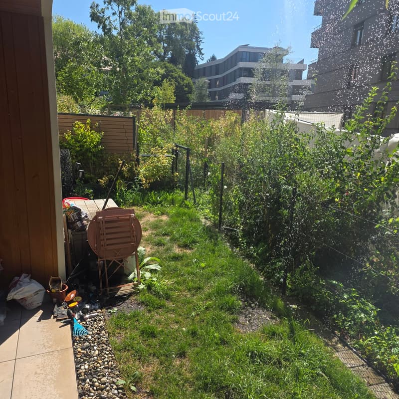 Garden area with lush greenery, a wooden fence, a small table with chairs, and a spray of water in the air.