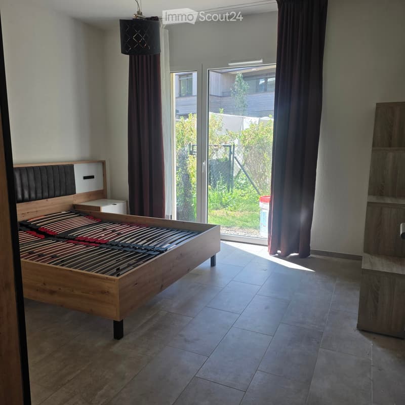 Modern bedroom with a wooden bed frame, nightstand, dark curtains, and a hanging lamp. The room has tiled flooring and large windows with a view of the garden.