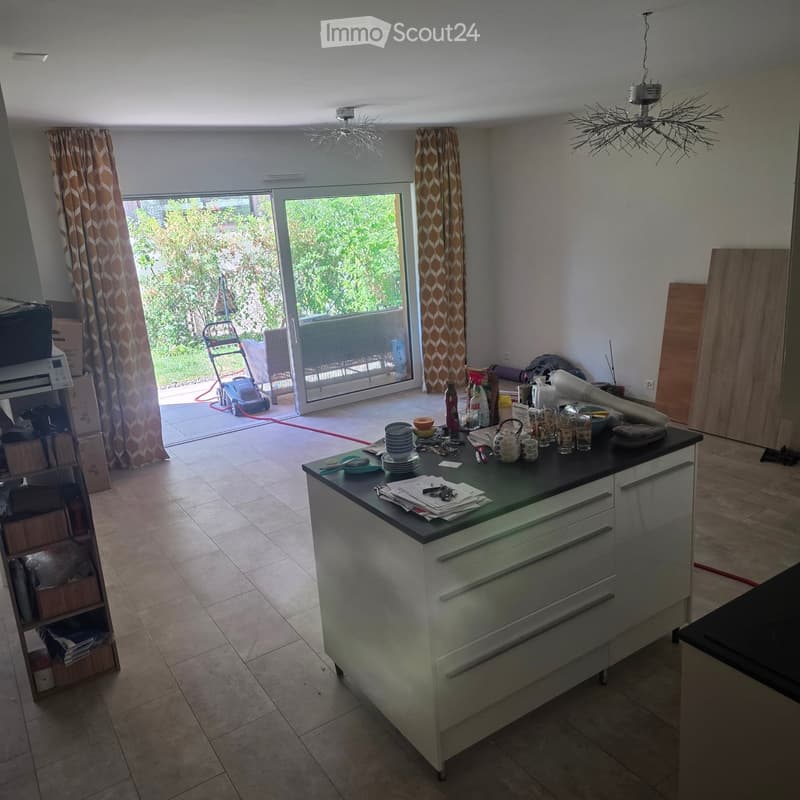 Modern kitchen with a central island, white cabinets, and a black countertop. The island has various items including cups, a teapot, and a bottle. There is a large sliding glass door leading to a patio with outdoor furniture and a lawn mower visible.