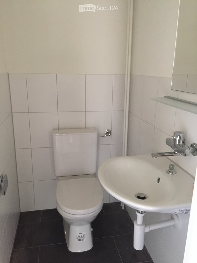 A small bathroom with a toilet, sink, and mirror. The walls are tiled in light beige, and the floor is dark gray.