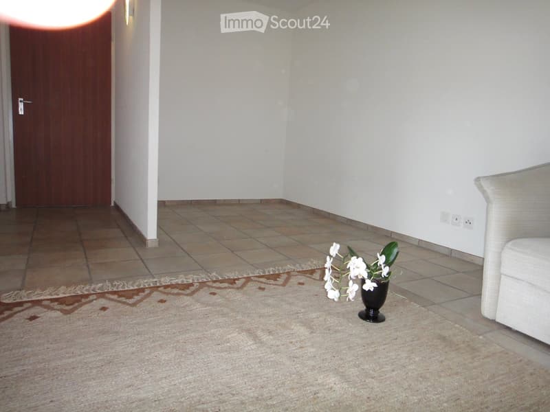 A small, well-lit room with tiled flooring, a patterned rug, a white sofa, and a vase with white flowers on the floor.