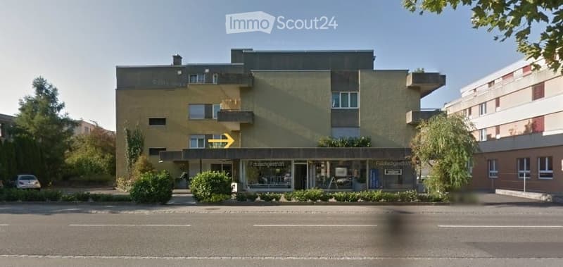 A multi-story building with a modern facade, featuring balconies and a storefront at the ground level. The building has a mix of light-colored walls and darker accents, with several windows visible.