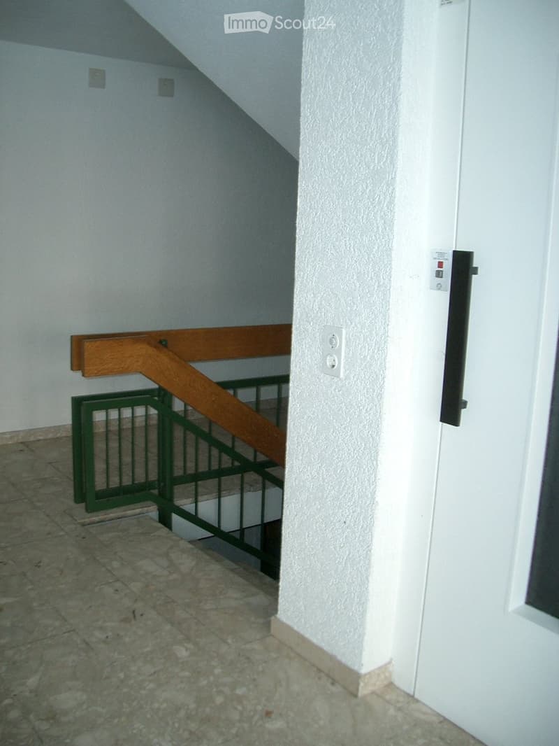 A narrow hallway with a staircase leading upwards, featuring a wooden handrail and green metal railing, light-colored tiled floor, and white walls.