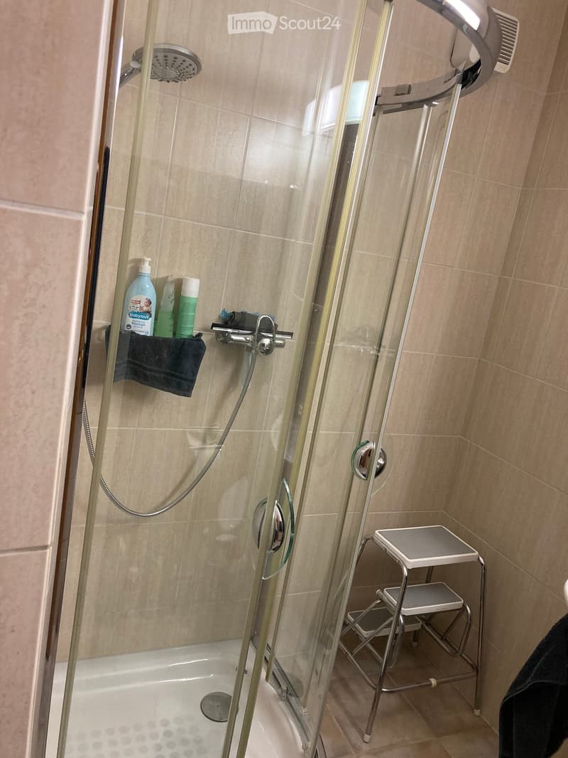 Modern bathroom with a bathtub, glass shower enclosure, and a small stool. The shower features a handheld sprayer and a fixed showerhead. A black towel and several bottles are placed on a shelf inside the shower.