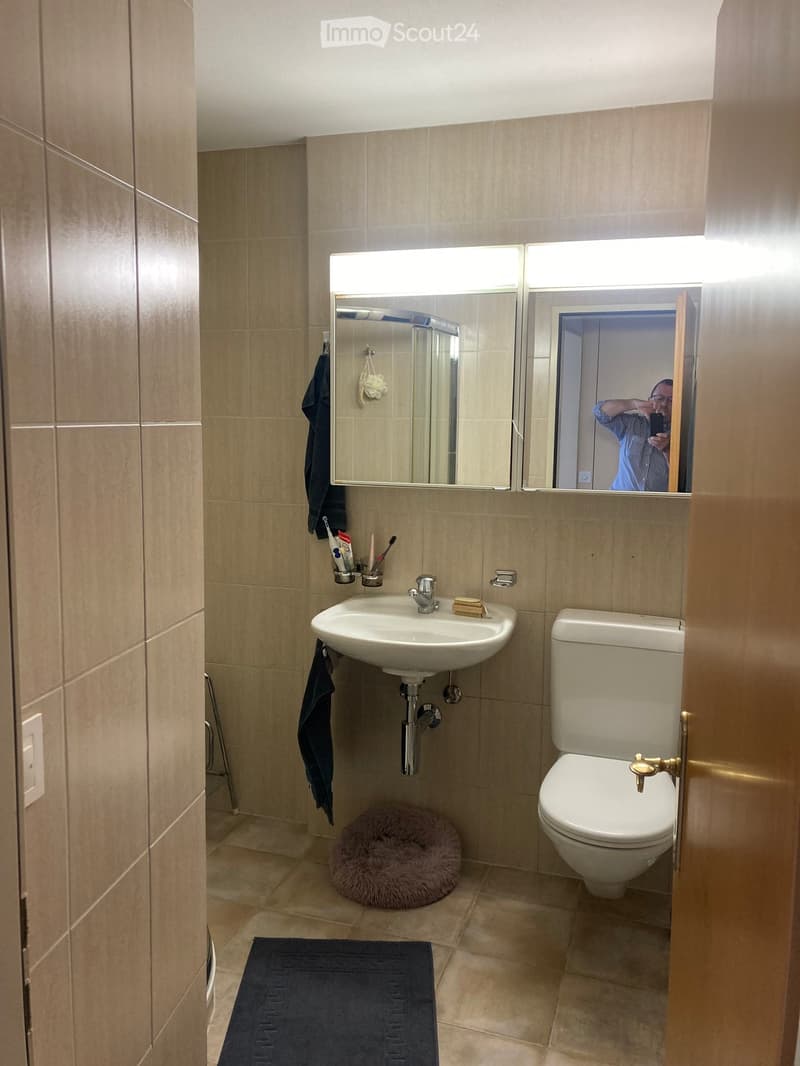Modern bathroom with beige tiles, a white sink, a toilet, a large mirror with built-in lighting, a towel, a small round stool, and a dark blue bath mat.