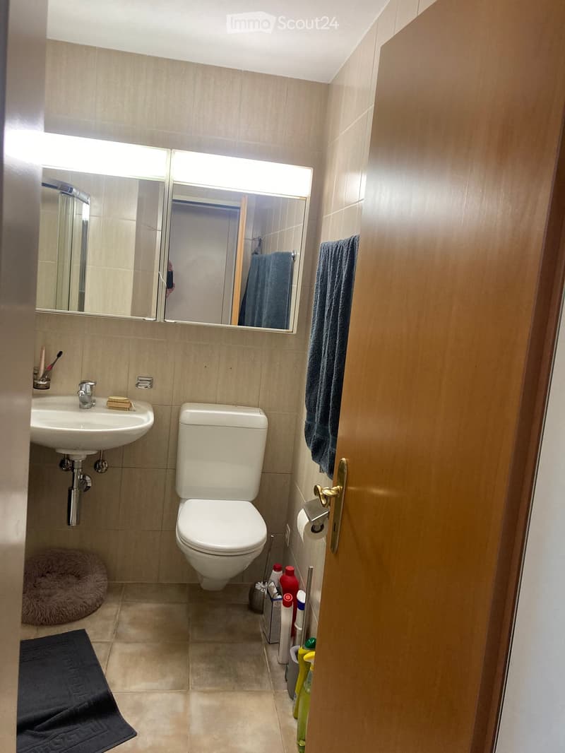 Modern bathroom with beige tiles, a toilet, a sink with a mirror above it, a towel on the wall, and cleaning supplies on the floor.