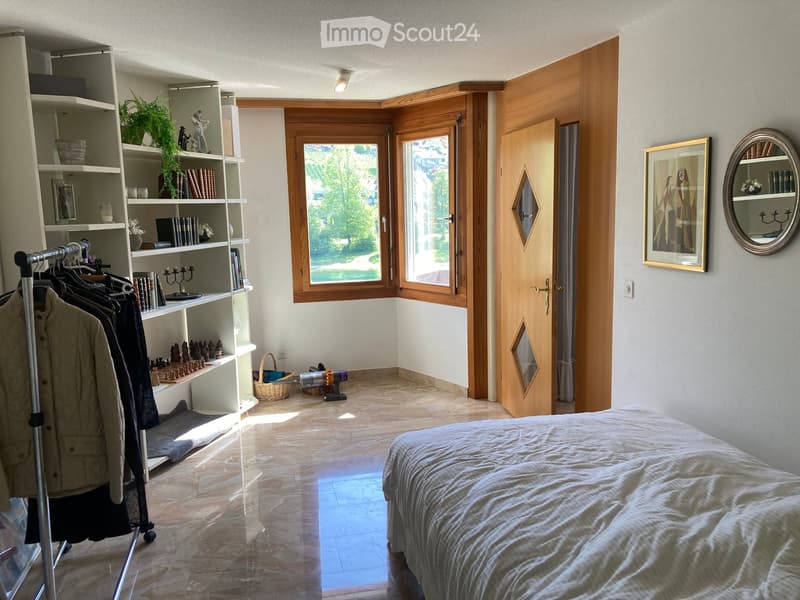 Bright bedroom with a large bed, wooden bookshelf, large window with view, and decorative items including a mirror and picture frame.