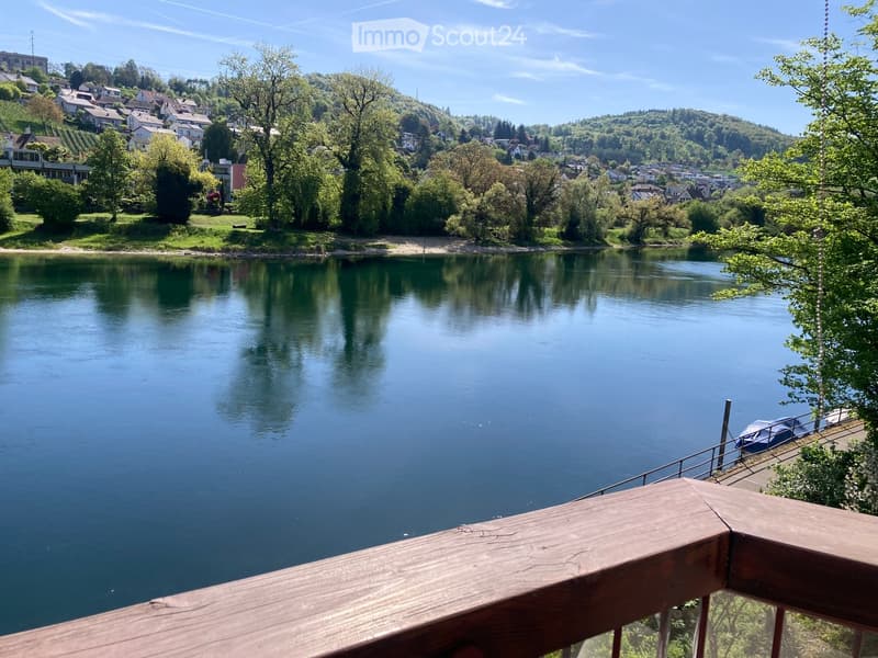 Balcony overlooking a serene river with a small dock and boat, surrounded by lush greenery and a town in the background.