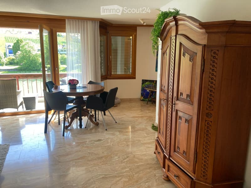 Dining area with a round wooden table, four black chairs, and a large wooden wardrobe. Large windows and sliding doors open to a balcony with outdoor seating.