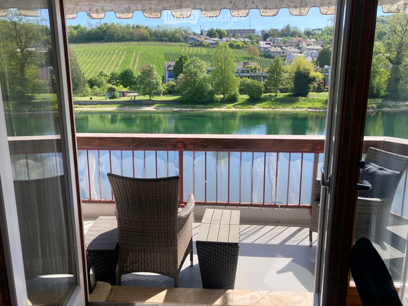 Balcony with wicker chairs and a small table, overlooking a scenic river and vineyard landscape.